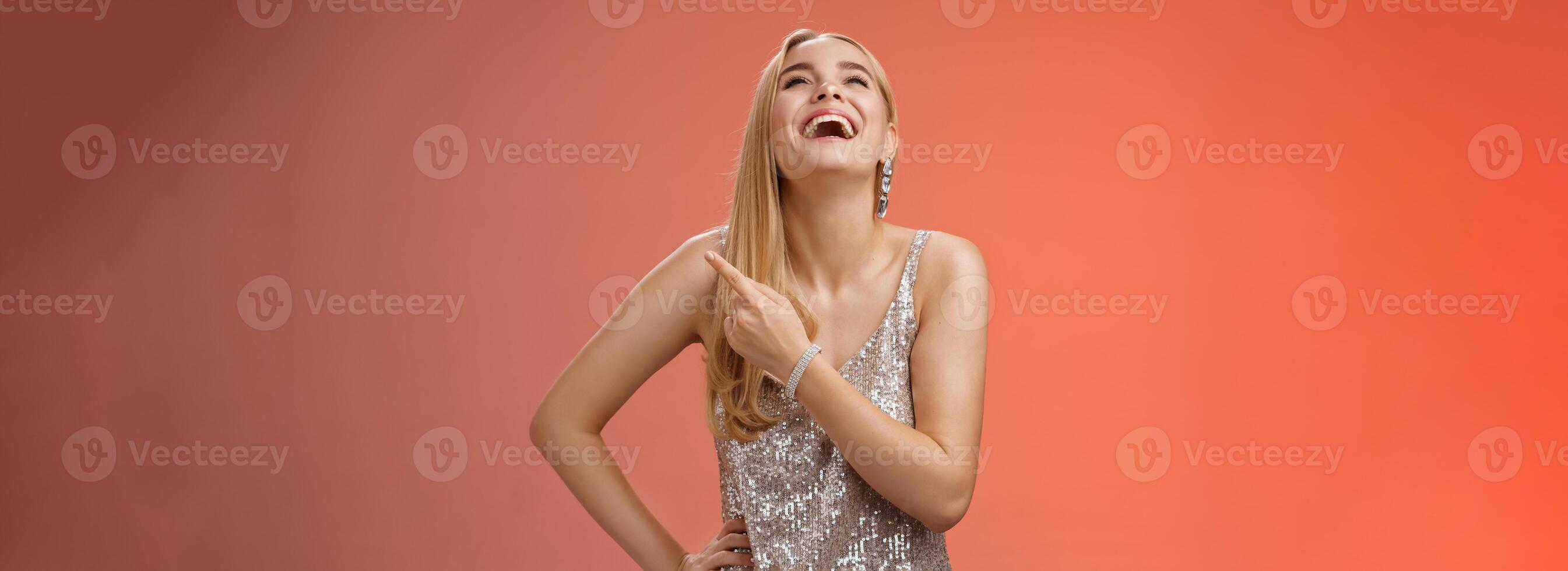 Fabulous carefree attractive blond woman in silver evening party dress laughing out loud have fun raise hand joyfully up pointing right index finger enjoying awesome sense humour, red background photo