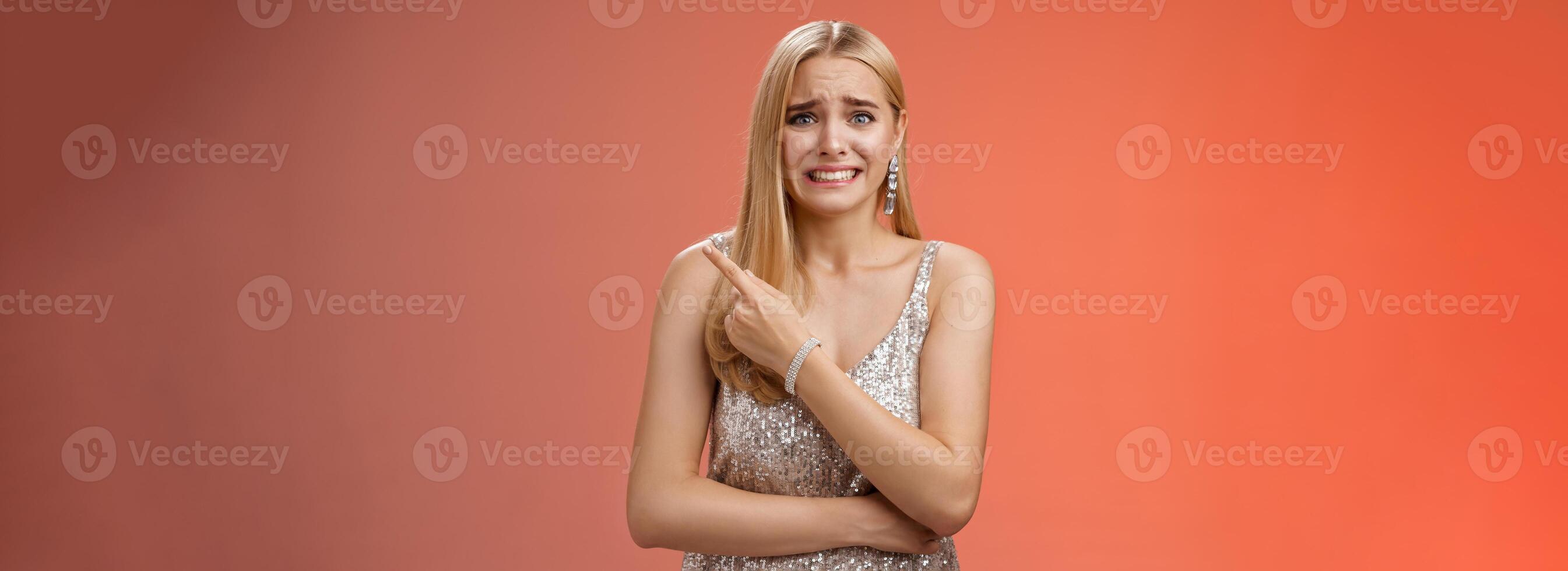 Nervous hesitant attractive worried blond woman cringing frowning clench teeth pointing behind seek friends help see ex-boyfriend unwilling be seen, standing anxious awkward party red background photo