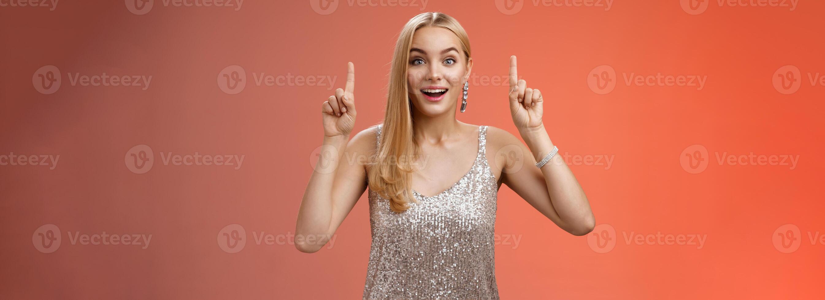 encantador asombrado rubio europeo mujer en fabuloso plata reluciente vestir aumento manos punto arriba divertido disfrutando acecho disparo estrellas, fuegos artificiales mirada cámara emocionado contento sorprendido, rojo antecedentes foto