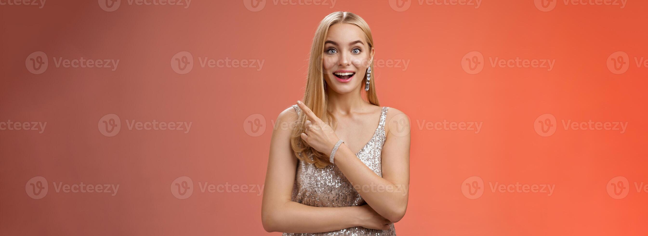 Joyful surprised good-looking blond woman talking friend during party smiling broadly look camera amused wondered pointing upper right corner curious discuss awesome room design, red background photo
