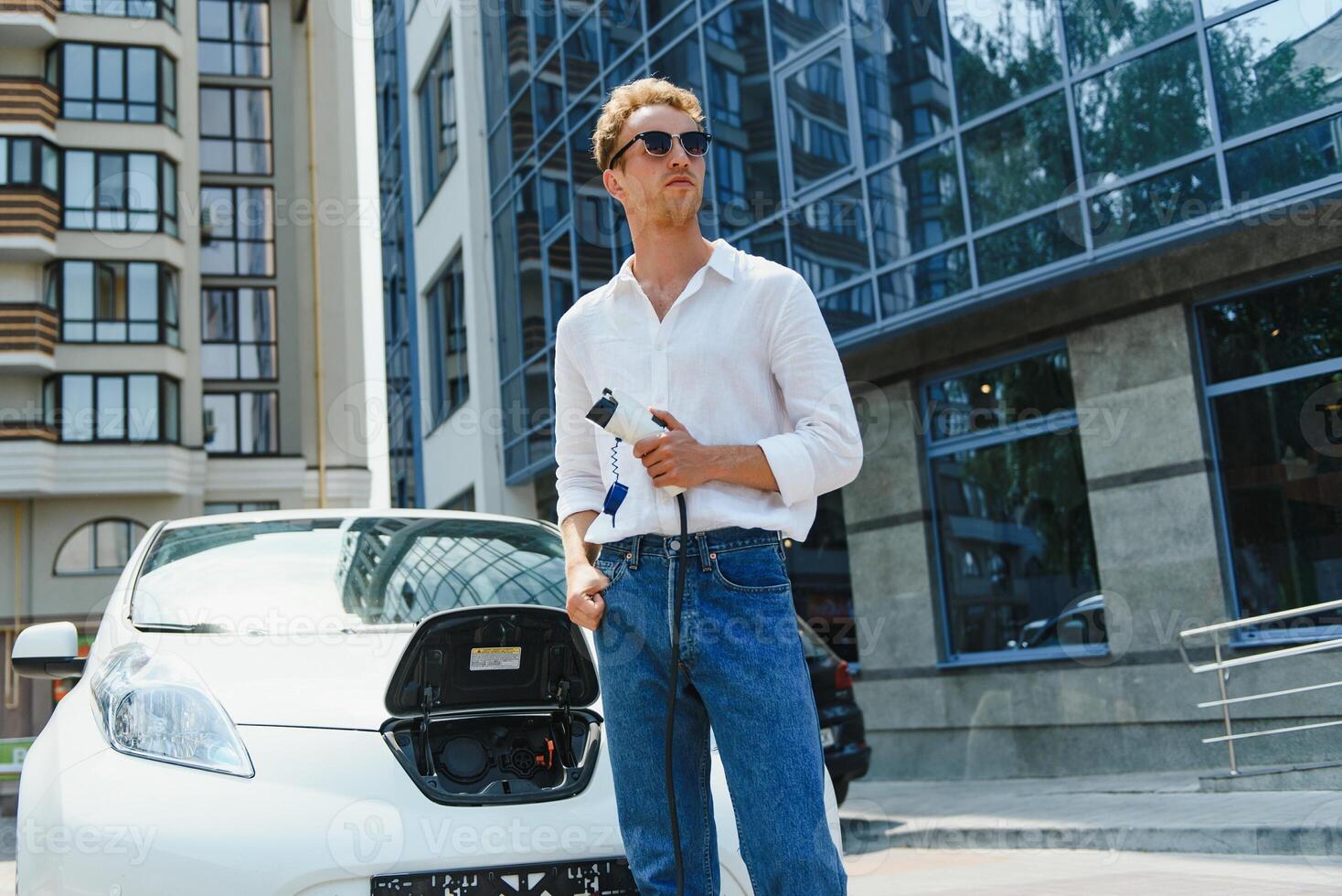 Man Holding Power Charging Cable For Electric Car In Outdoor Car Park. And he s going to connect the car to the charging station in the parking lot near the shopping center photo