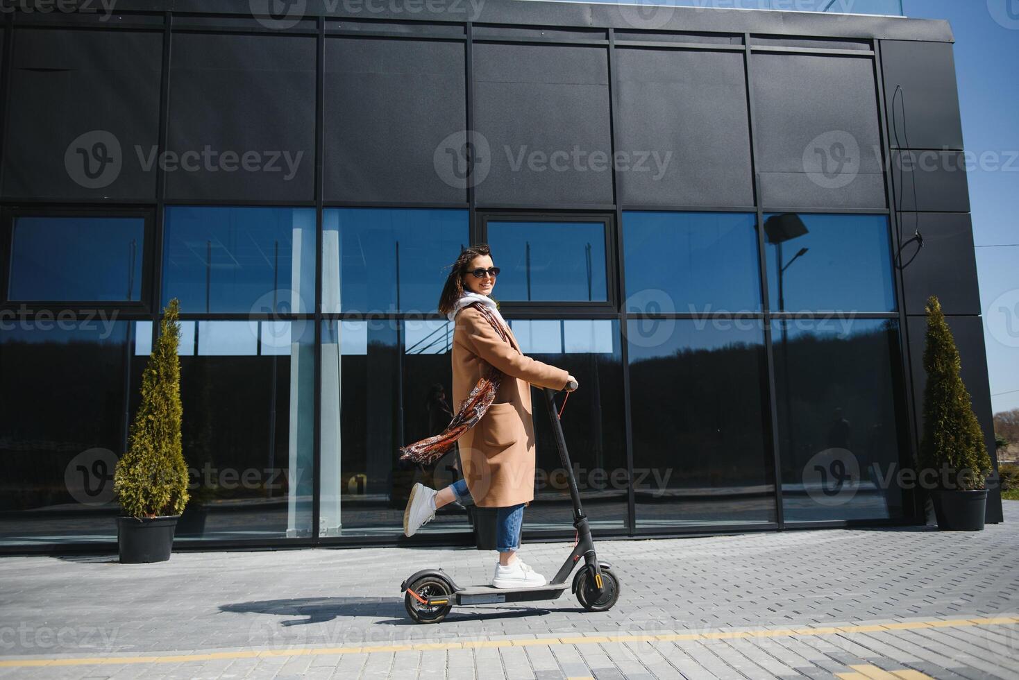 joven hermosa mujer en un chaqueta sonrisas y paseos un eléctrico scooter a trabajo a lo largo oficina edificios foto