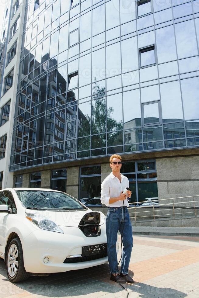 Stylish man with coffe cup in hand inserts plug into the electric car charging socket photo