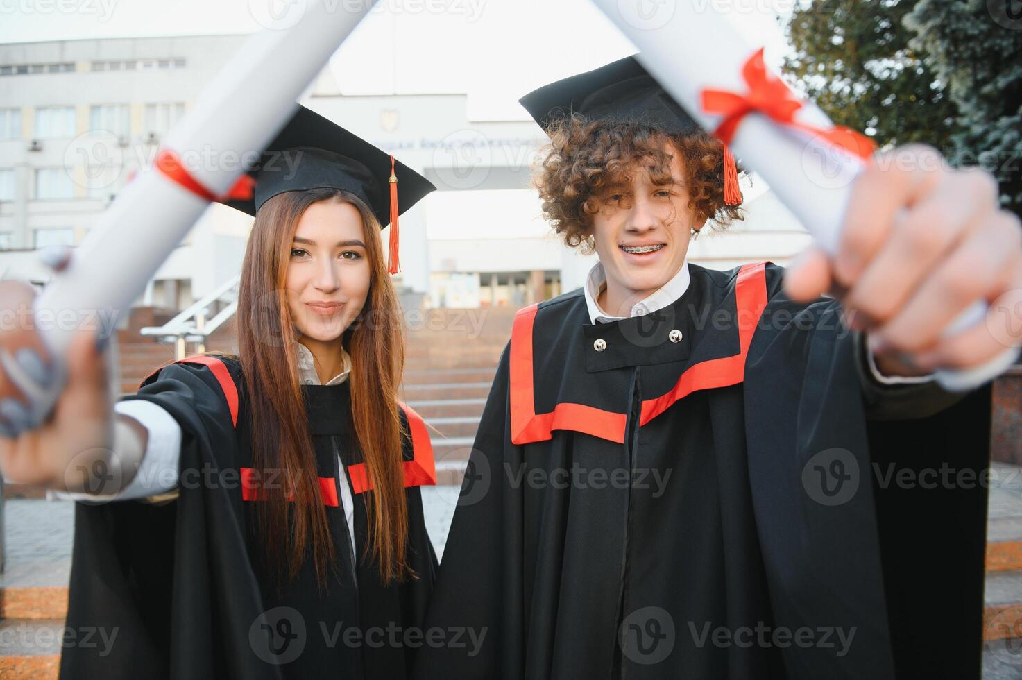 retrato de contento graduados dos amigos en graduación tapas y vestidos en pie fuera de Universidad edificio con otro estudiantes en fondo, participación diploma pergaminos, y sonriente foto