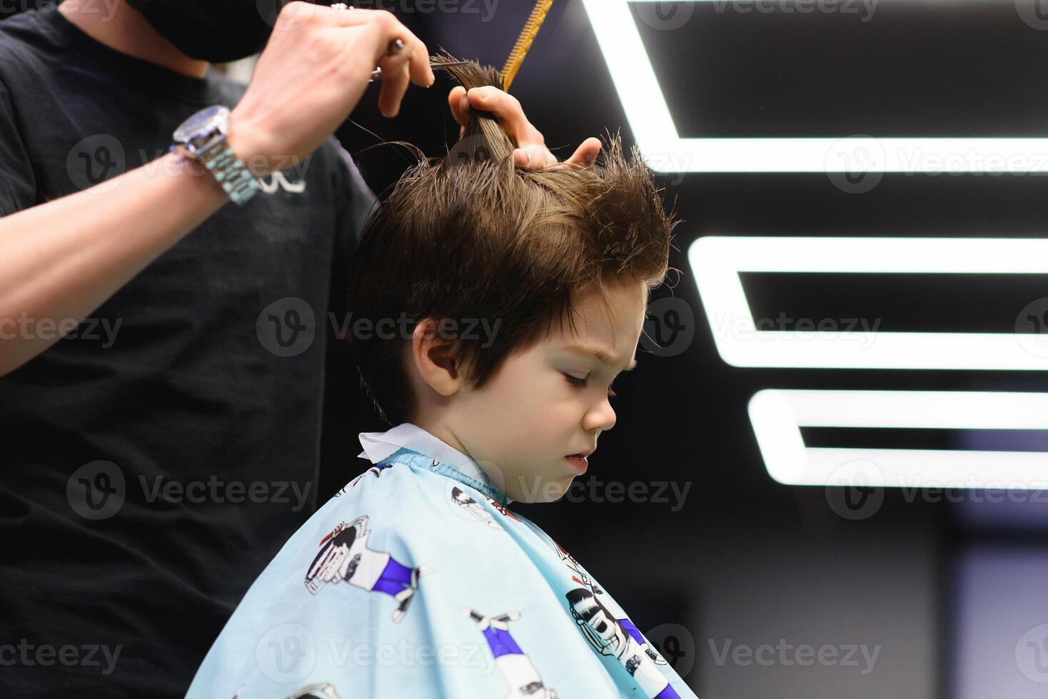 Side view of cute little boy getting haircut by hairdresser at the barbershop. photo