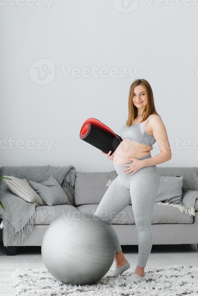 retrato de un hermosa embarazada mujer sonriente y participación un arrollado yoga estera debajo su brazo. adorable expectante madre conmovedor su barriga y preparando a comienzo su hogar rutina de ejercicio foto