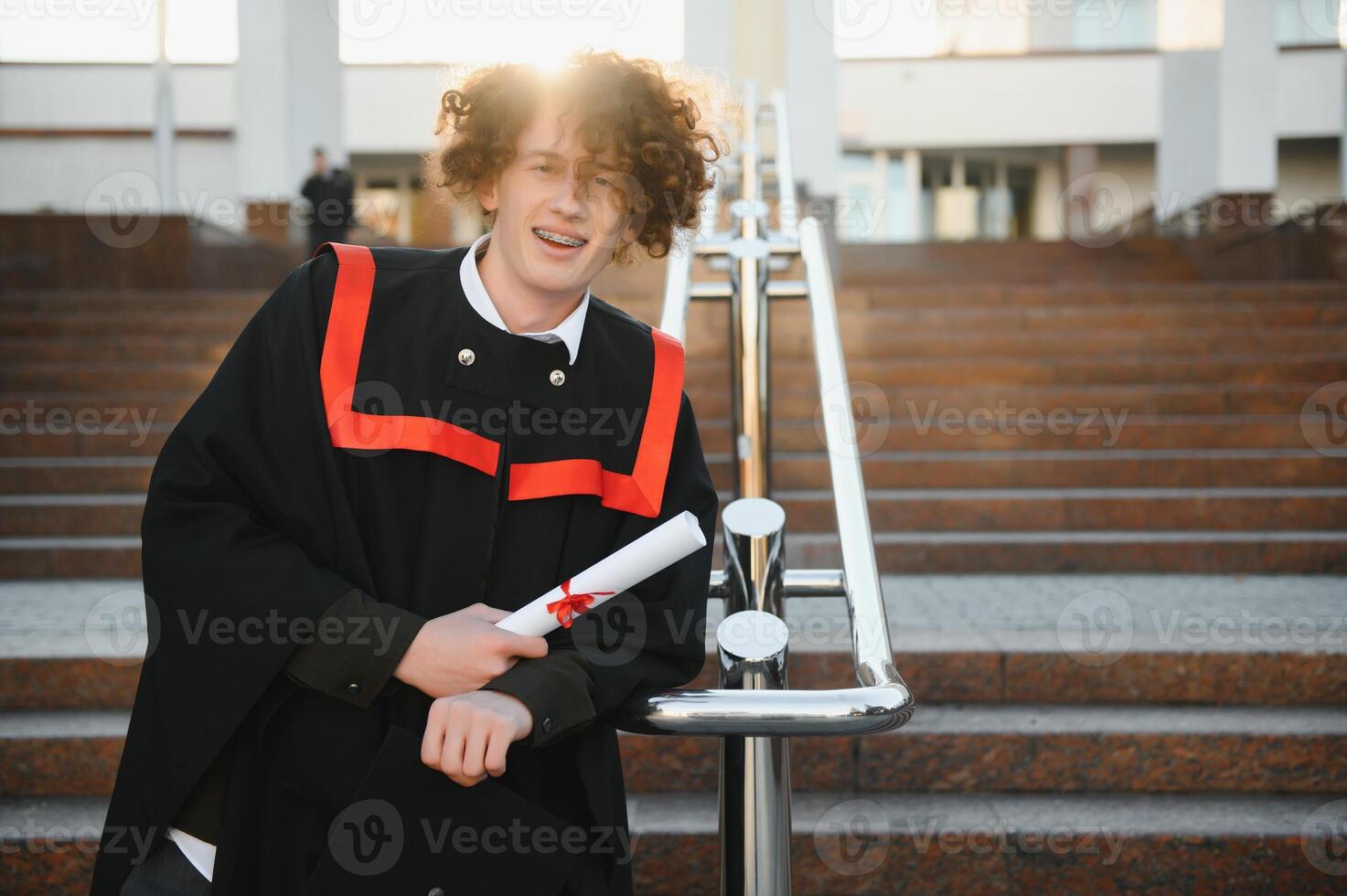 Graduation from university. Young smiling boy university graduate in traditional bonet and mantle standing and holding diploma in hand over university building background photo
