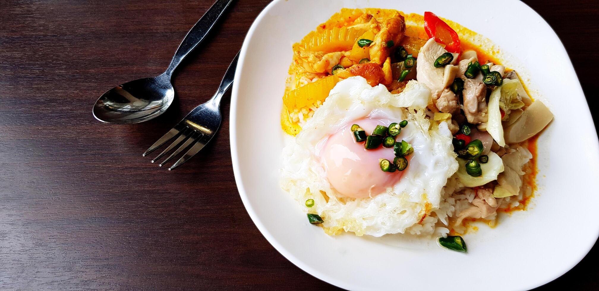 Hot Yellow curry with pork, vegetable boiled, Fried egg on top with rice and stainless steel spoon and fork in white plate on dark brown wooden table with copy space. Thai curry food concept photo