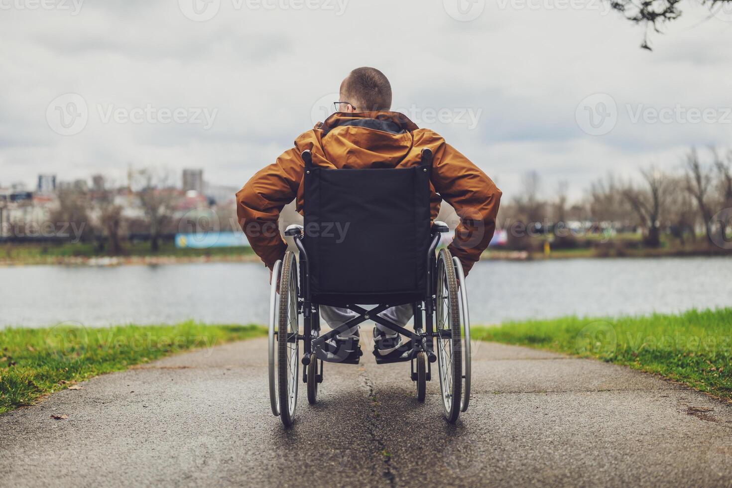 Rear view image of paraplegic handicapped man in wheelchair by the lake. He is rolling on walkway. photo