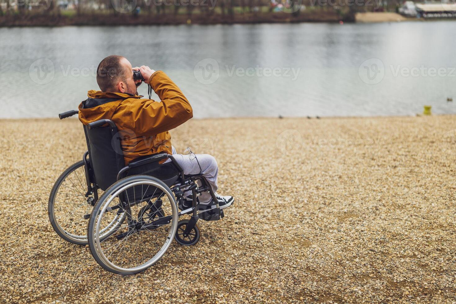 Paraplegic handicapped man in wheelchair is using binoculars outdoor. He is watching nature by the lake. photo