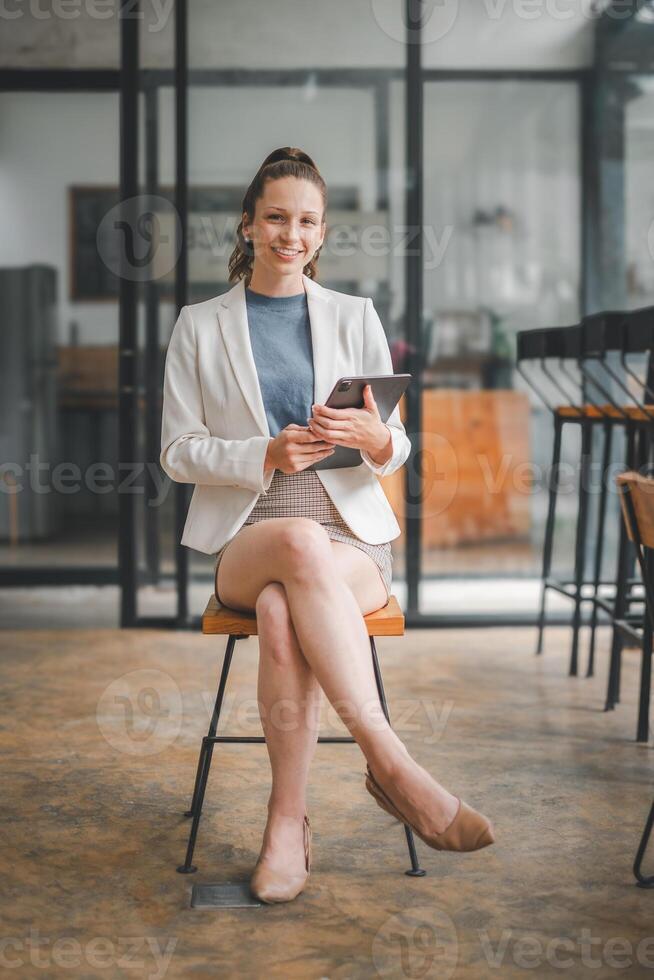 Business analytics concept, Elegant businesswoman in a modern office setting sits on a high stool, holding a digital tablet with a pleasant smile. photo