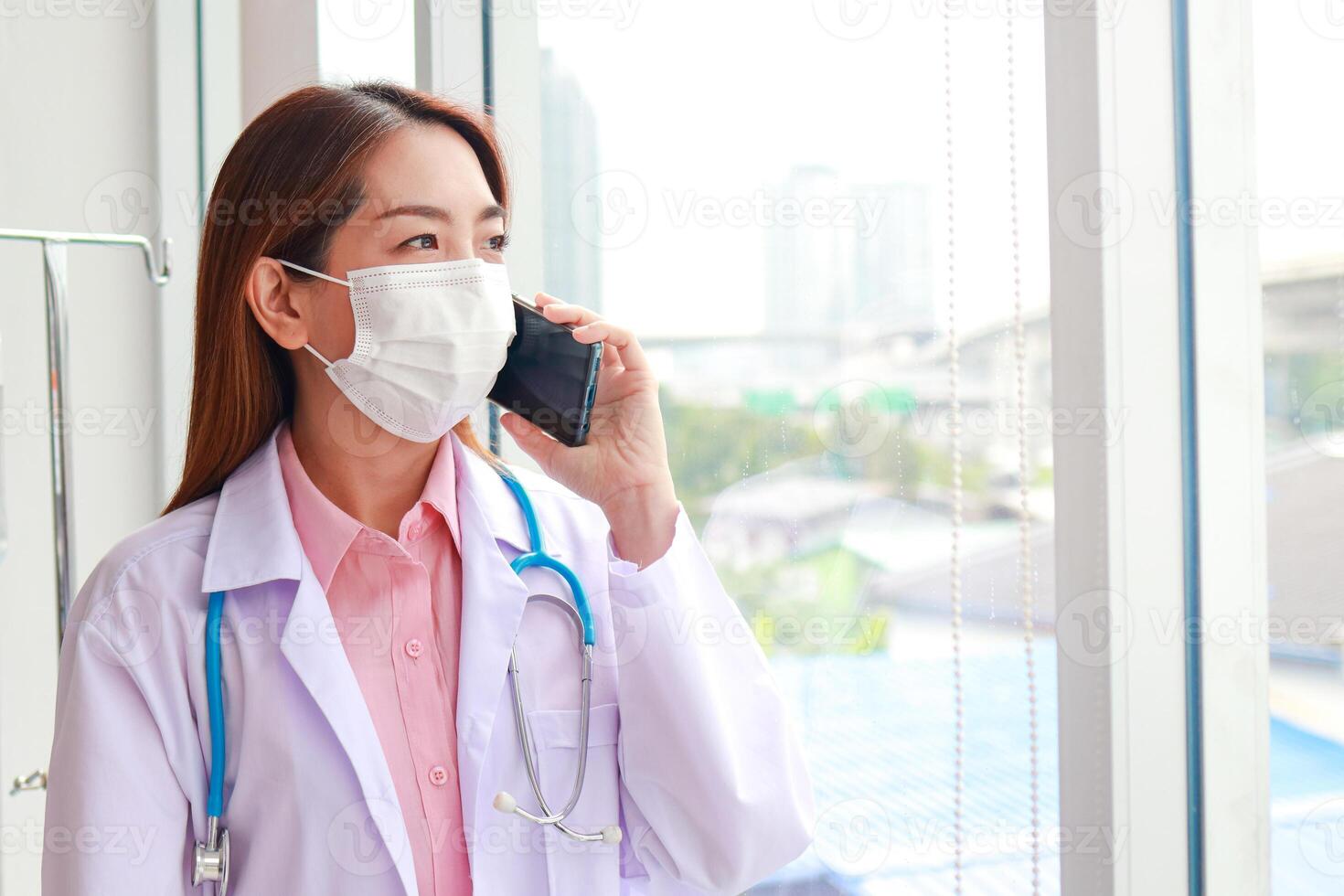hermosa asiático hembra médico vistiendo un blanco máscara sostener un teléfono inteligente y hablar a el paciente. moderno comunicación tecnología concepto, paciente cuidado mediante teléfono inteligente Copiar espacio foto