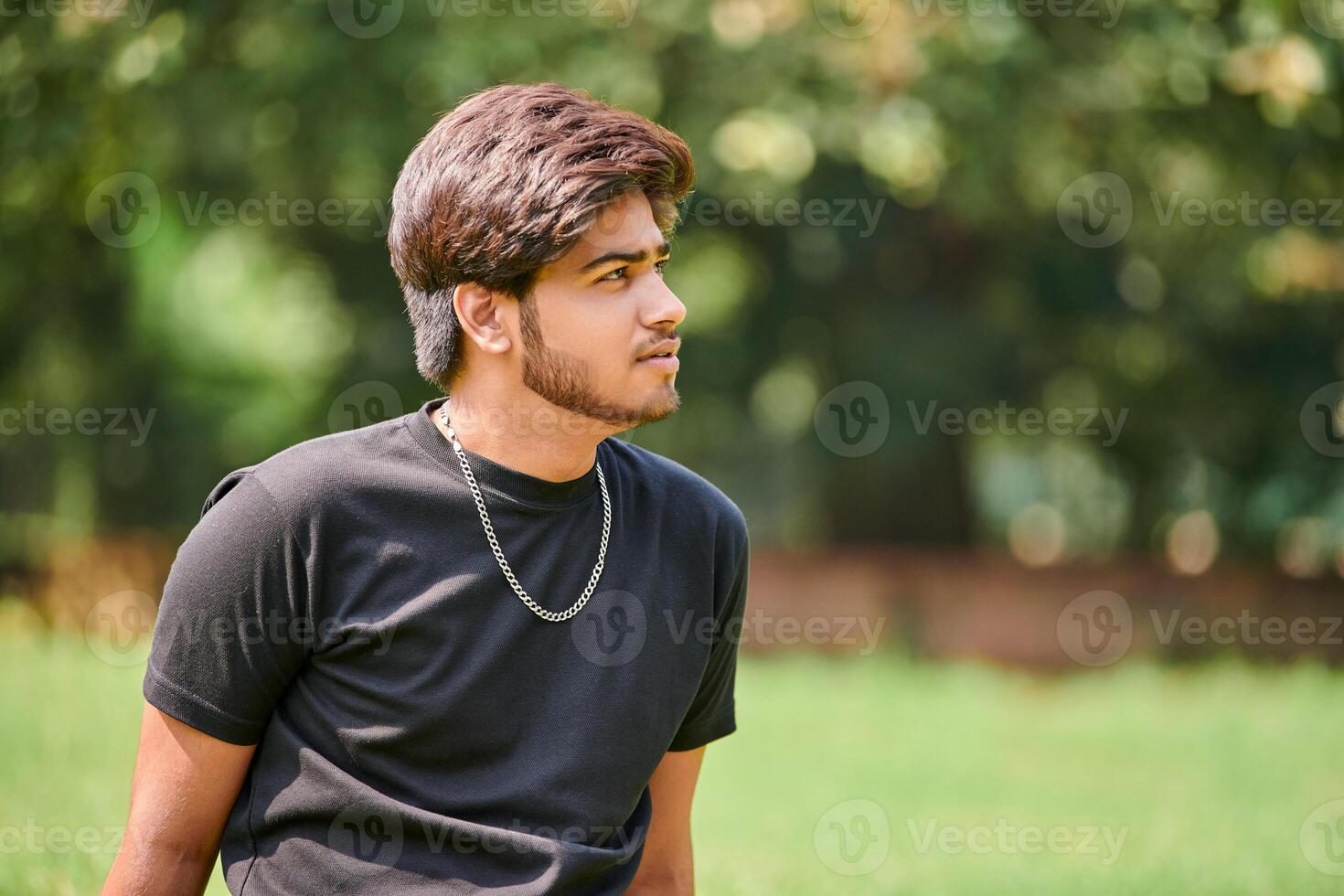 Attractive young indian man portrait in black t shirt and silver neck chain sitting on green lawn photo