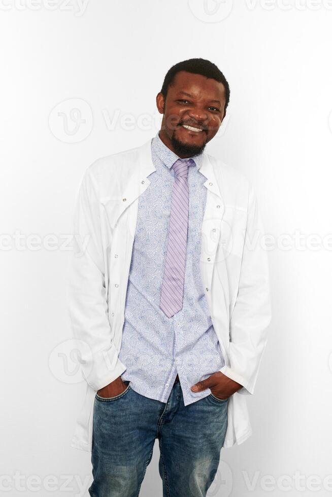 Happy black doctor man with small beard in white coat, bright shirt, tie and jeans, white background photo