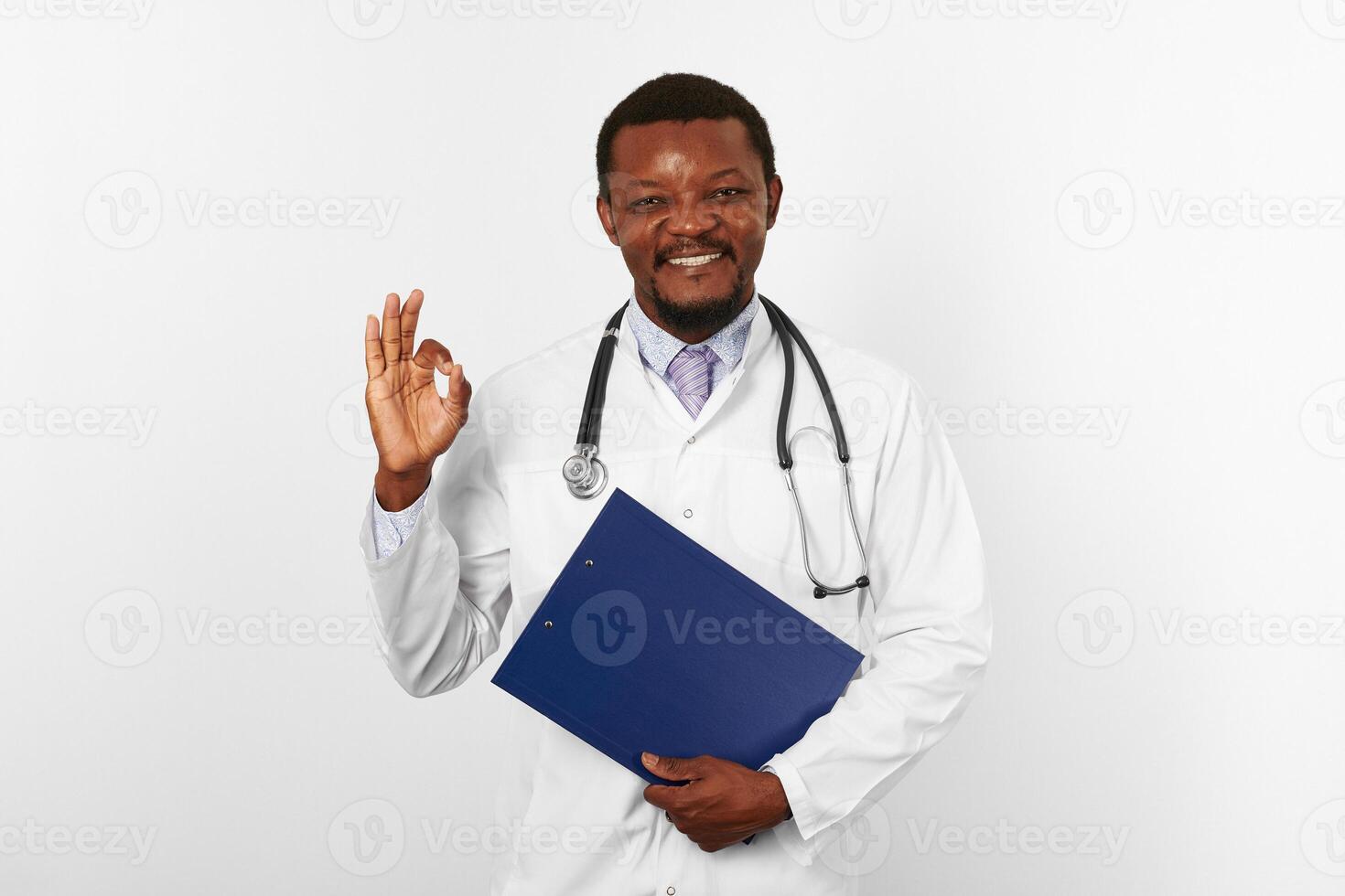 Smiling black bearded doctor man in white robe holds medical clipboard and shows OK gesture photo