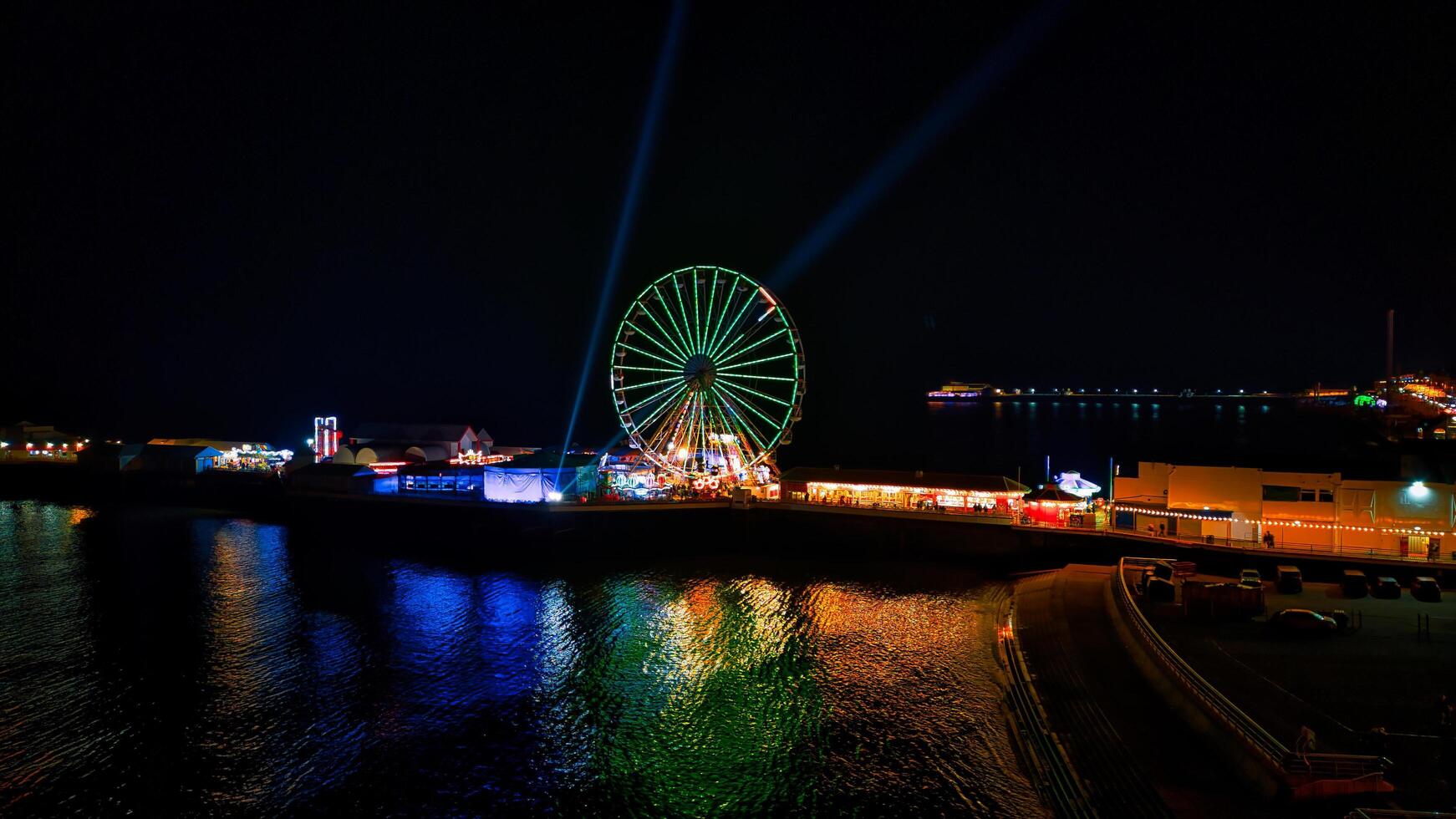 Nighttime view of a lit Ferris wheel by the water with colorful reflections and searchlights in the sky in Backpool, England. photo