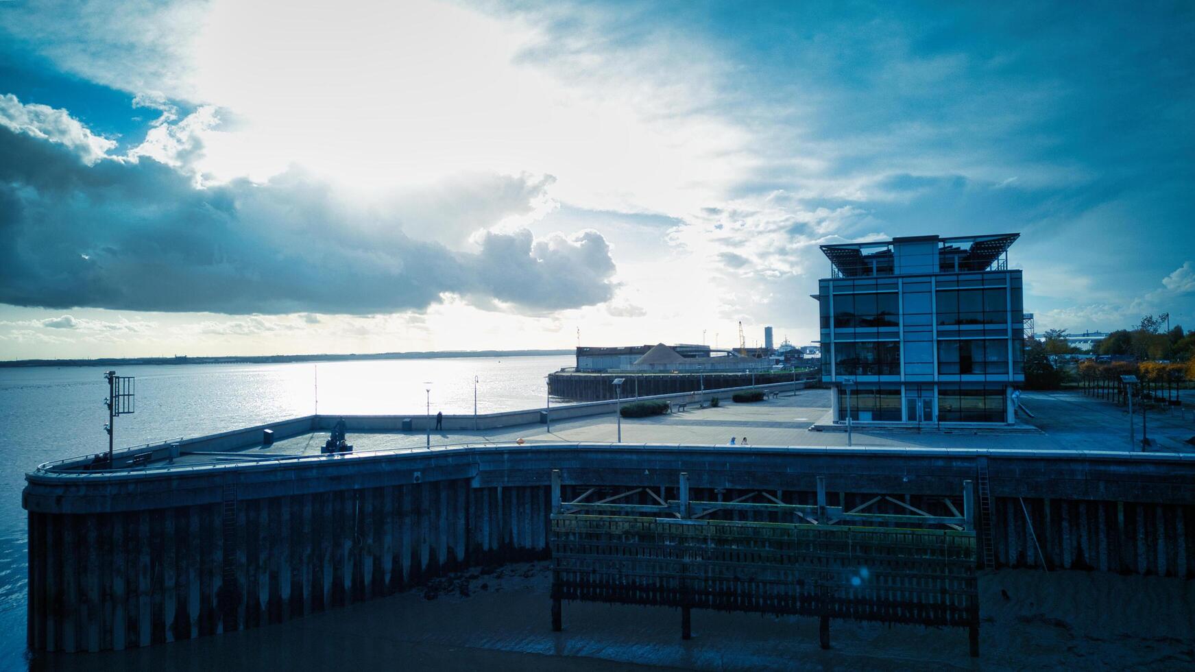 Dramatic cloudscape over a modern waterfront building with a serene river view and a silhouette of a railing in the foreground in Hull, England. photo