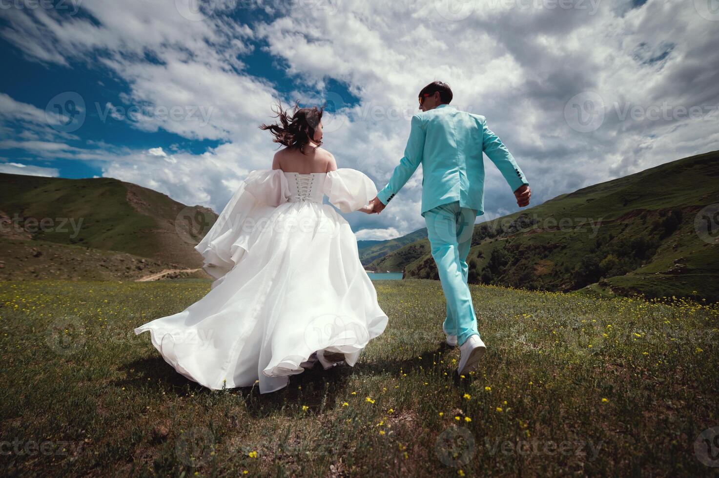 Rear view of running loving newlyweds holding hands, a bride in a wedding dress and a wife in a turquoise suit running across a field against the backdrop of green hills photo