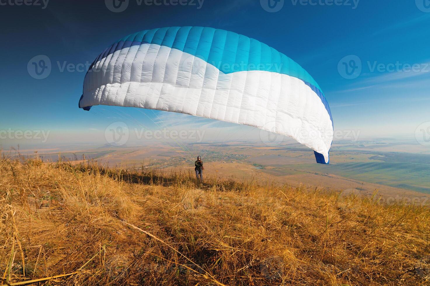 A paraglider takes off from a mountainside with a blue and white canopy and the sun behind. A paraglider is a silhouette. The glider is sharp, with little wing movement. photo