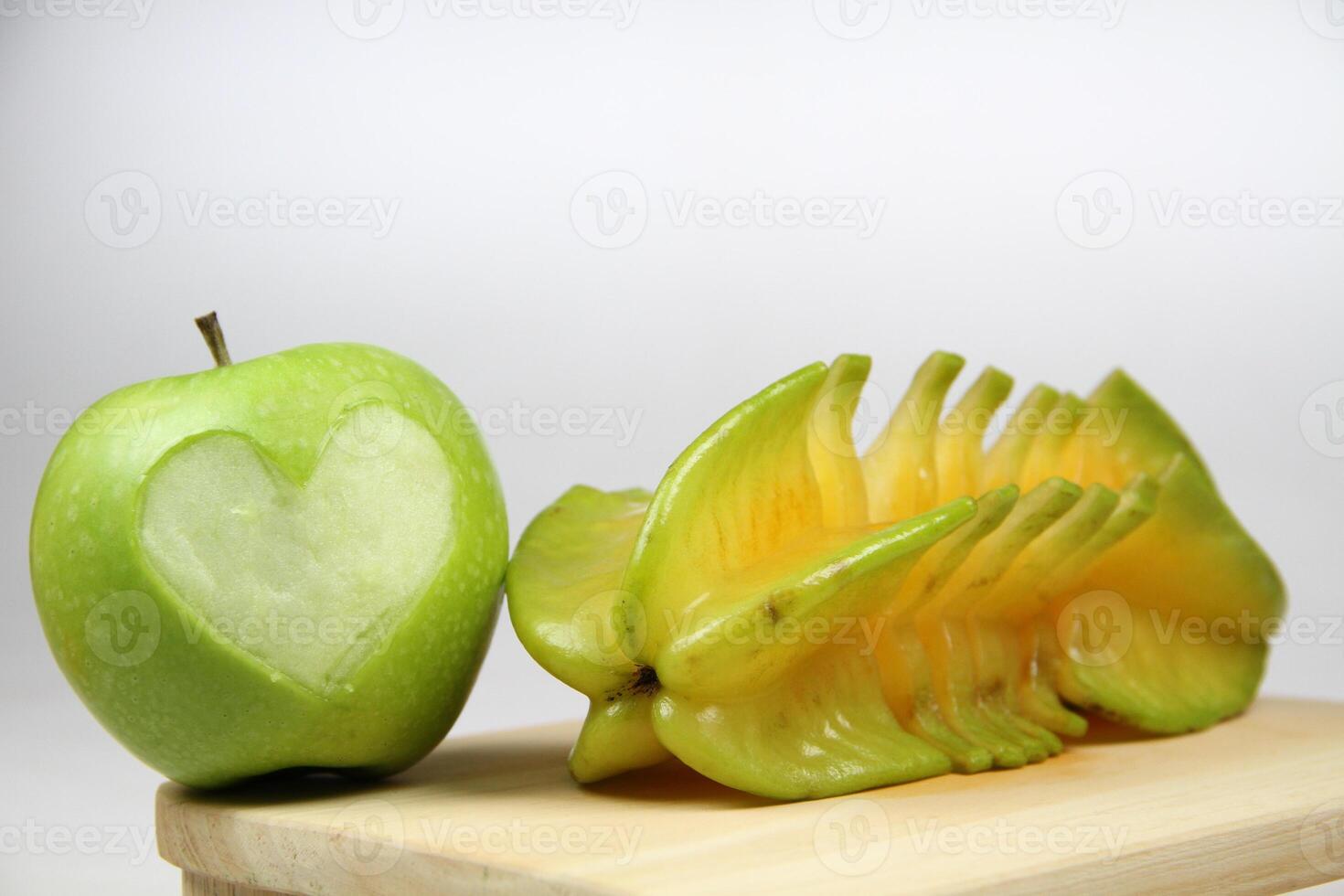 a green apple shaped like a heart and slices of starfruits well arranged on wooden cutting board on white background photo
