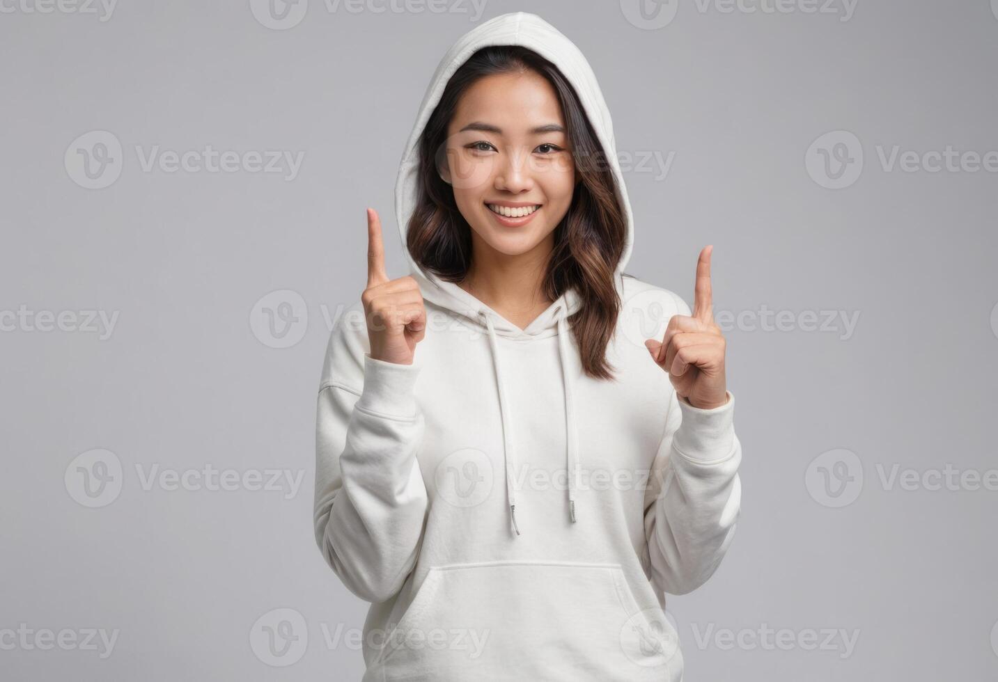 AI Generated An Asian woman in a white hoodie points upwards with both hands, showing enthusiasm and a bright smile. photo