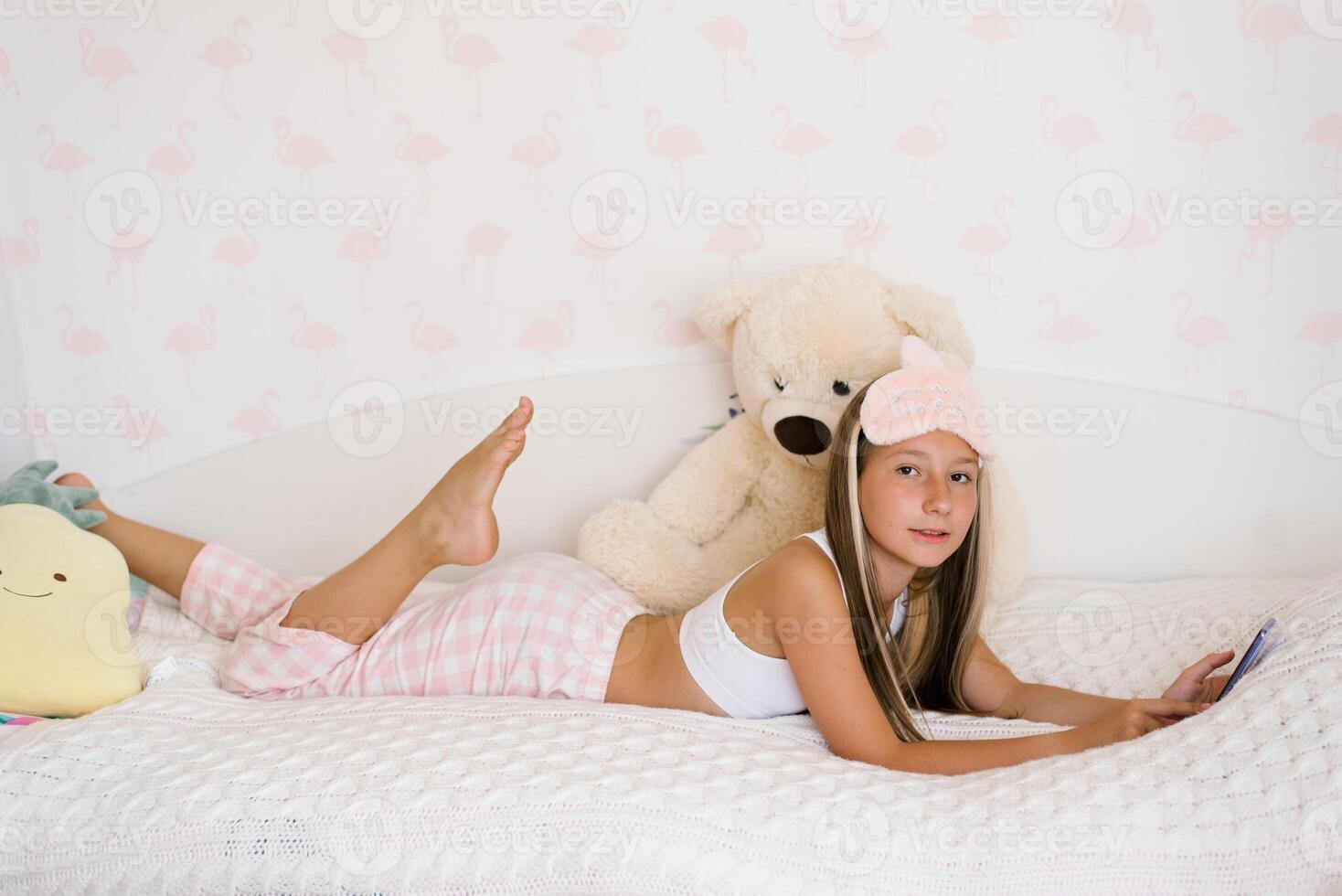 Beautiful teenage girl in pajamas is lying on a cozy bed on her stomach and surfing on the phone in the house in the morning, the younger generation is addicted to gadgets photo