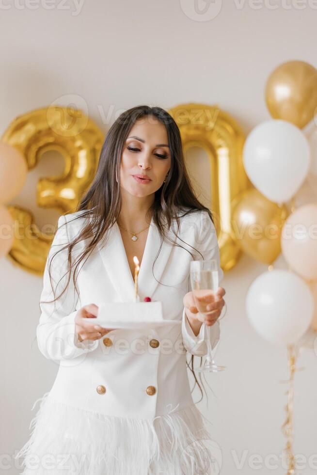 Positive woman celebrating anniversary dressed in stylish feather dress blowing out candle on birthday cake around inflated balloons at home photo