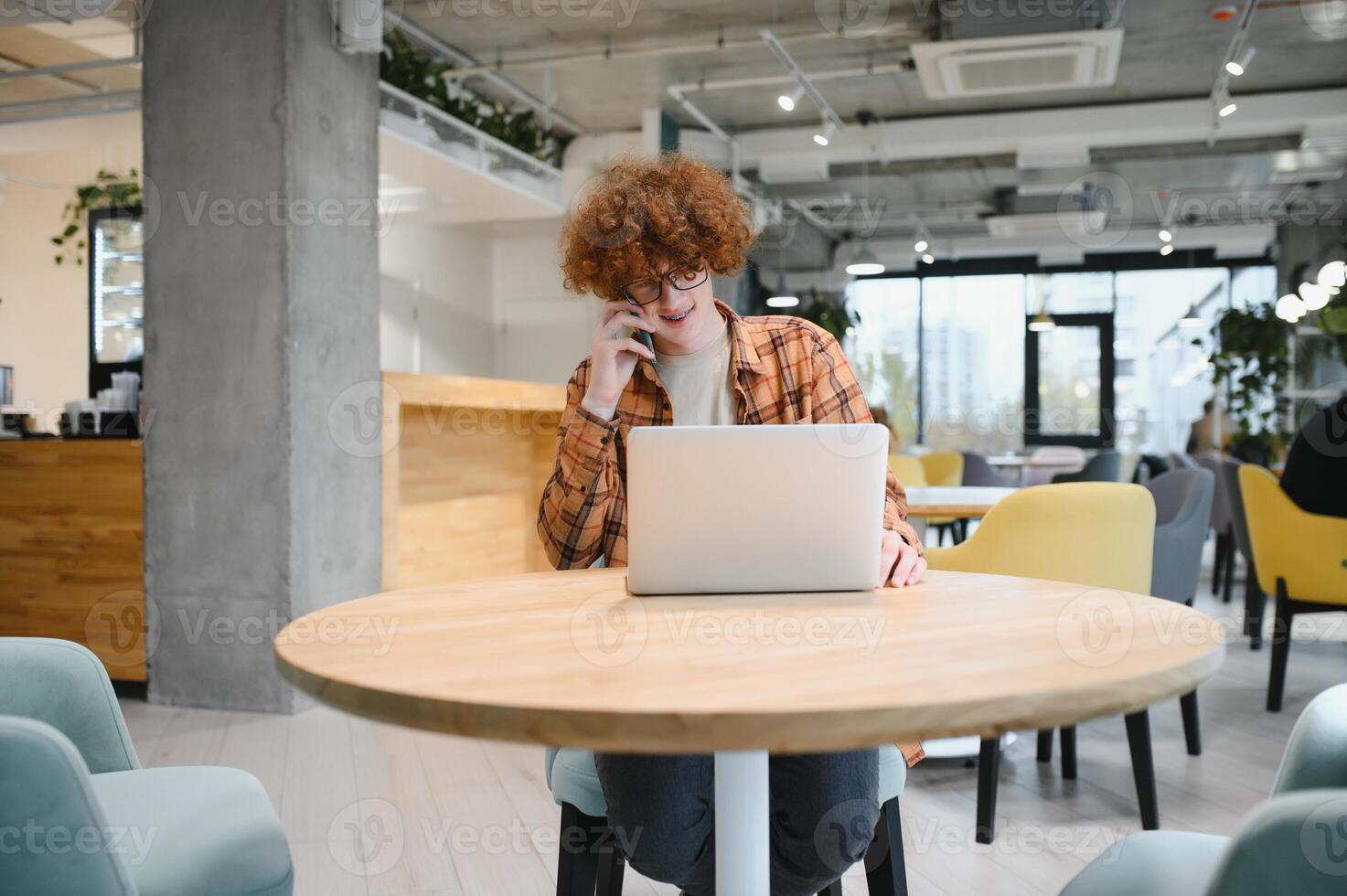 Young happy male freelancer in casual clothes sitting in cafe with laptop and using mobile phone. photo