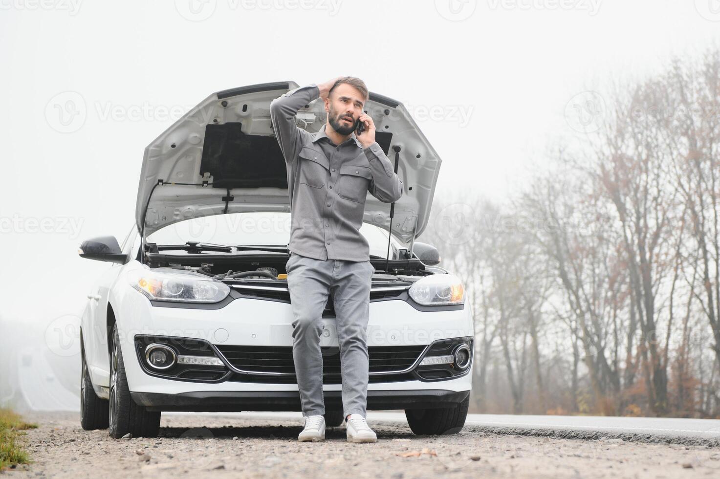 A young man near a broken car with an open hood on the roadside. photo