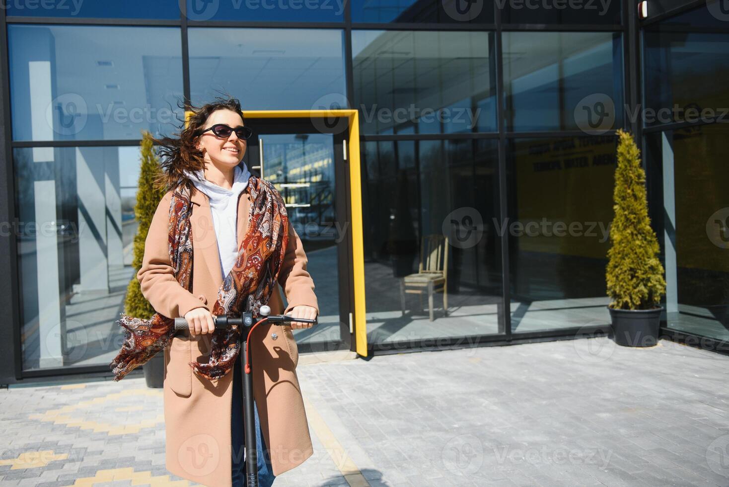 Young beautiful woman in a jacket smiles and rides an electric scooter to work along office buildings photo