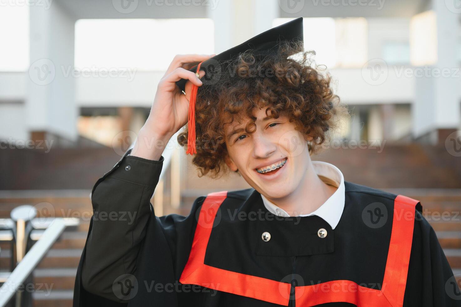 Graduation from university. Young smiling boy university graduate in traditional bonet and mantle standing and holding diploma in hand over university building background photo