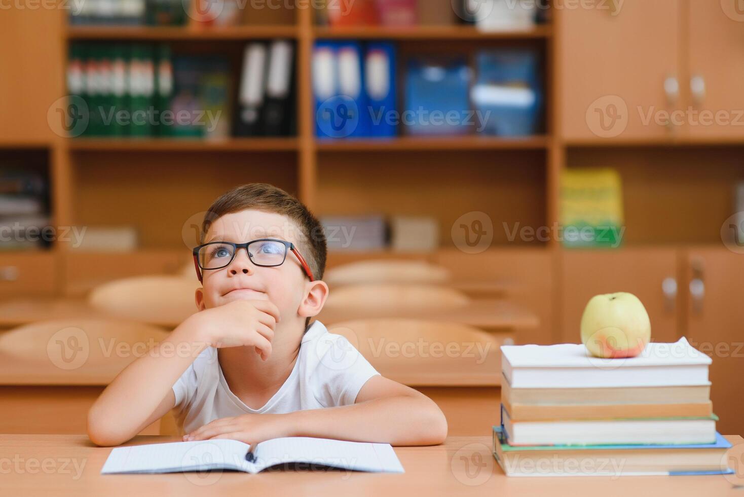 Concentrated schoolboy sitting at desk and writing in exercise book with classmate sitting behind photo