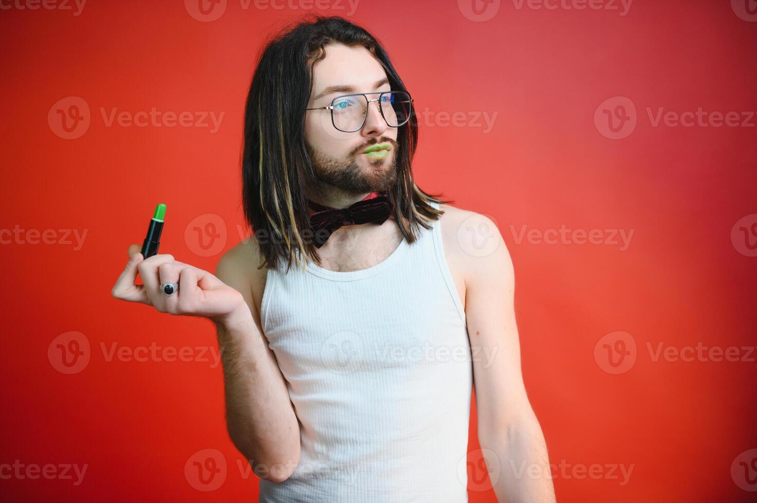 Young man applying lip gloss on his lips and looking in camera like mirror. Gay man doing make up against red background photo