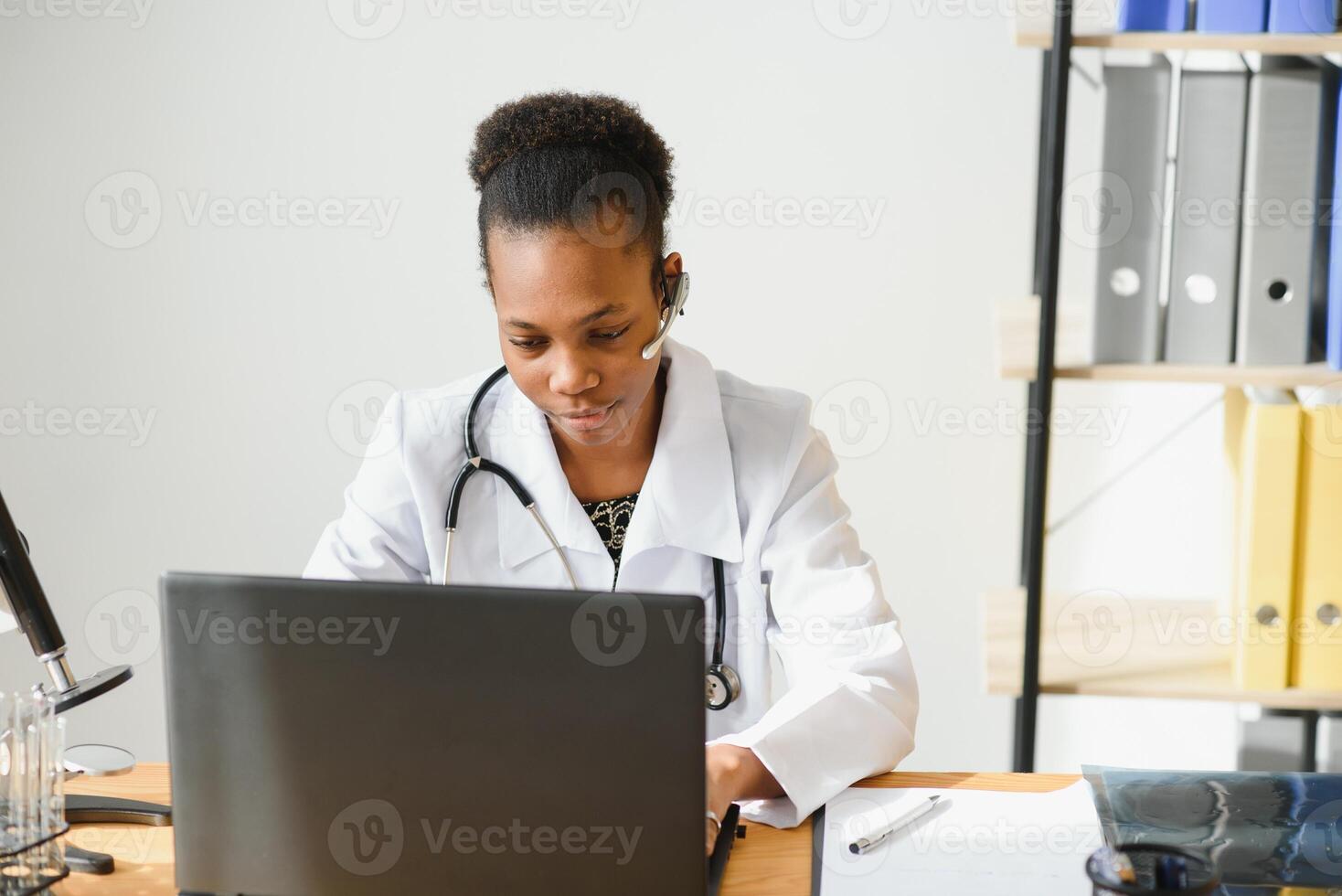 Smiling african american female doctor gp wears white medical coat using laptop computer at workplace gives remote online consultation, working on pc, consulting patient in internet telemedicine chat photo