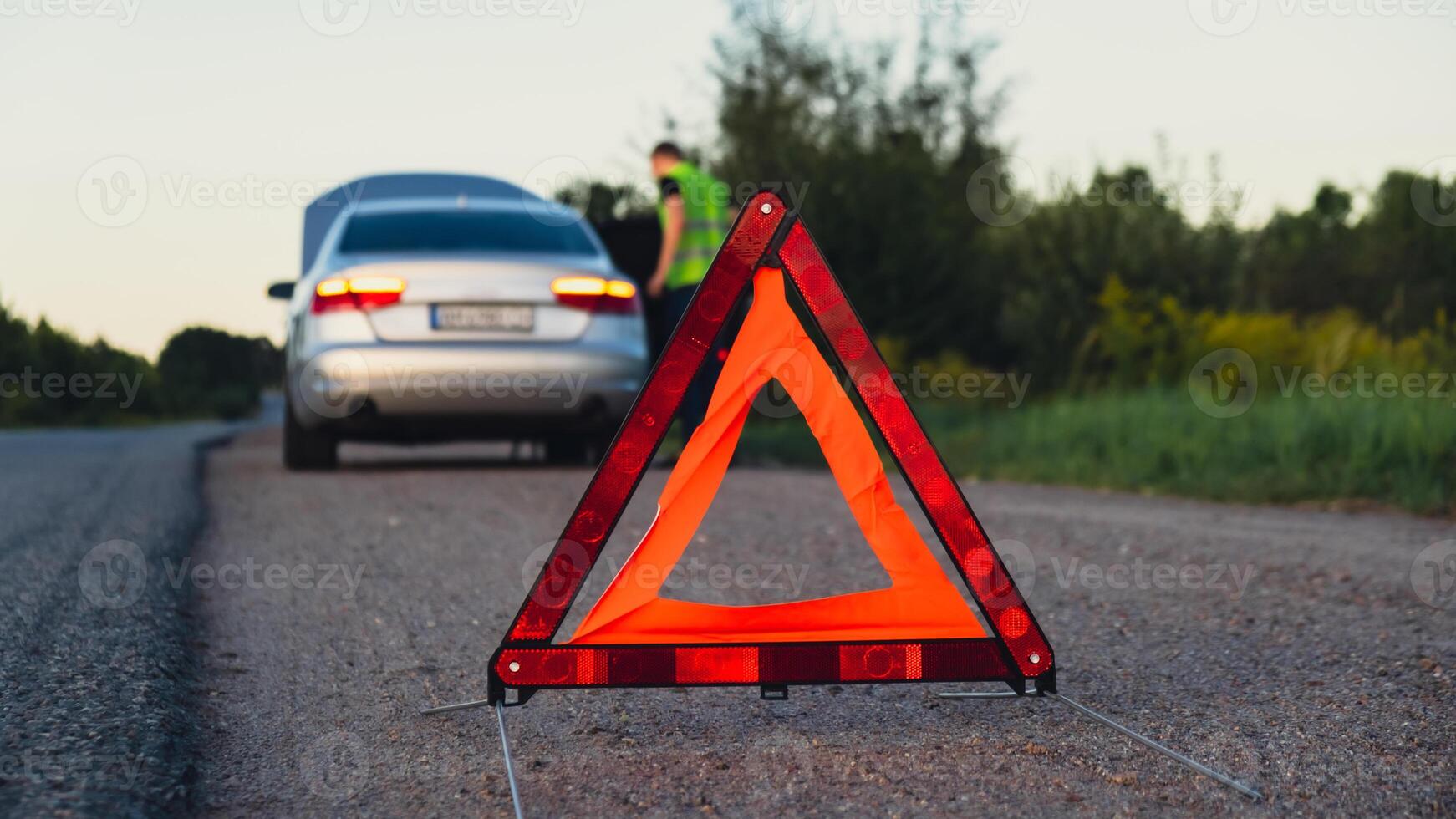 Broken silver luxury car emergency accident. Man driver installing red triangle stop sign on road. Sport automobile turned on blinkers technical problems on the road. Safety procedure when having vehicle broken down photo