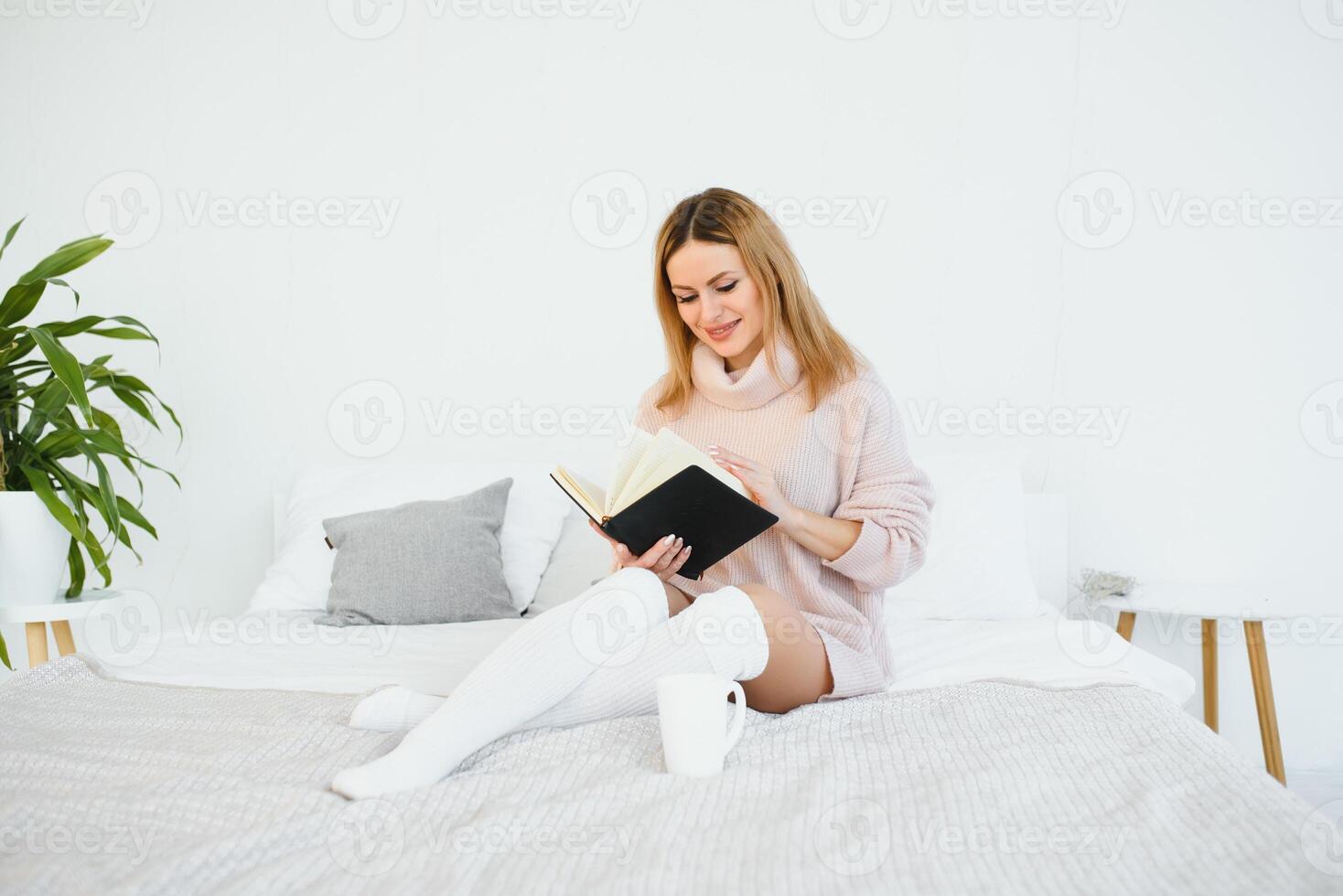 A woman reading a book and smiling as she sits in bed. The alarm clock on the desk beside her. photo