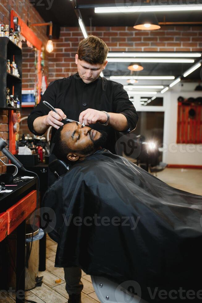 lado ver de grave hombre con elegante moderno Corte de pelo mirando adelante en Barbero tienda. mano de Barbero acuerdo Derecho maquinilla de afeitar y corte de moda rayas en cabeza de cliente. foto