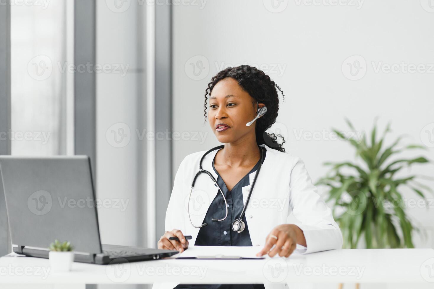 medicine, online service and healthcare concept - happy smiling african american female doctor or nurse with headset and laptop having conference or video call at hospital. photo