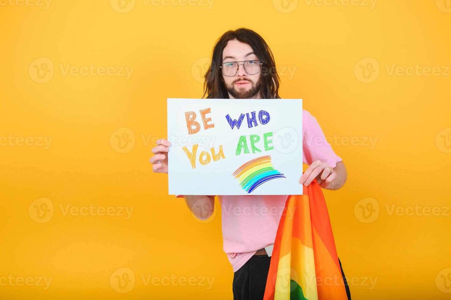 Young queer bisexual smiling gay man with make up in beige tank shirt hold card sign with be who you are title text on rainbow flag background studio portrait. photo