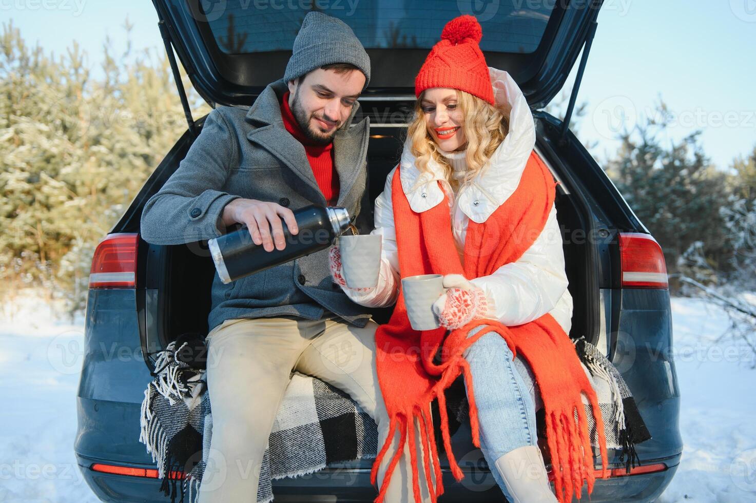 Couple in love sitting in car trunk drinking hot tea in snowy winter forest and chatting. People relaxing outdoors during road trip. Valentines day photo