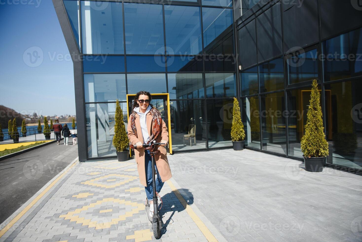 joven hermosa mujer en un chaqueta sonrisas y paseos un eléctrico scooter a trabajo a lo largo oficina edificios foto