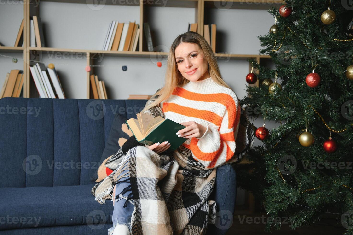 Attractive young lady with dark hair reading interesting book while sitting on grey couch. Blur background of beautiful christmas tree. Cozy atmosphere. photo