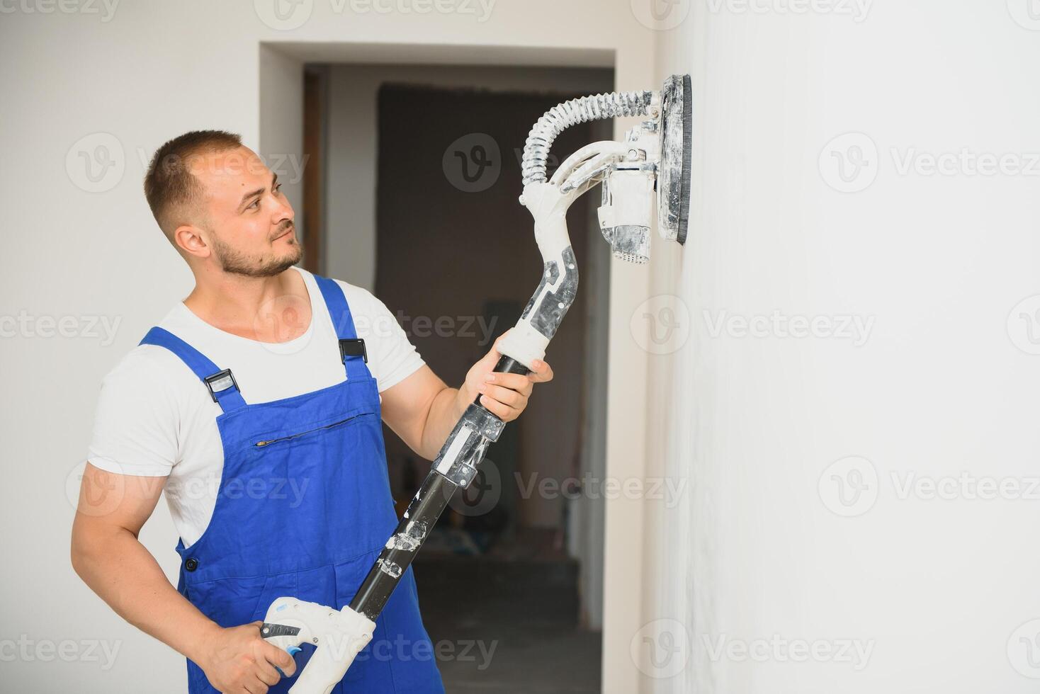 Plasterer smoothes the wall surface with a wall grinder. Master builder grind a white plaster wall. a man in overalls grinds the surface in a respirator. experienced repairman photo