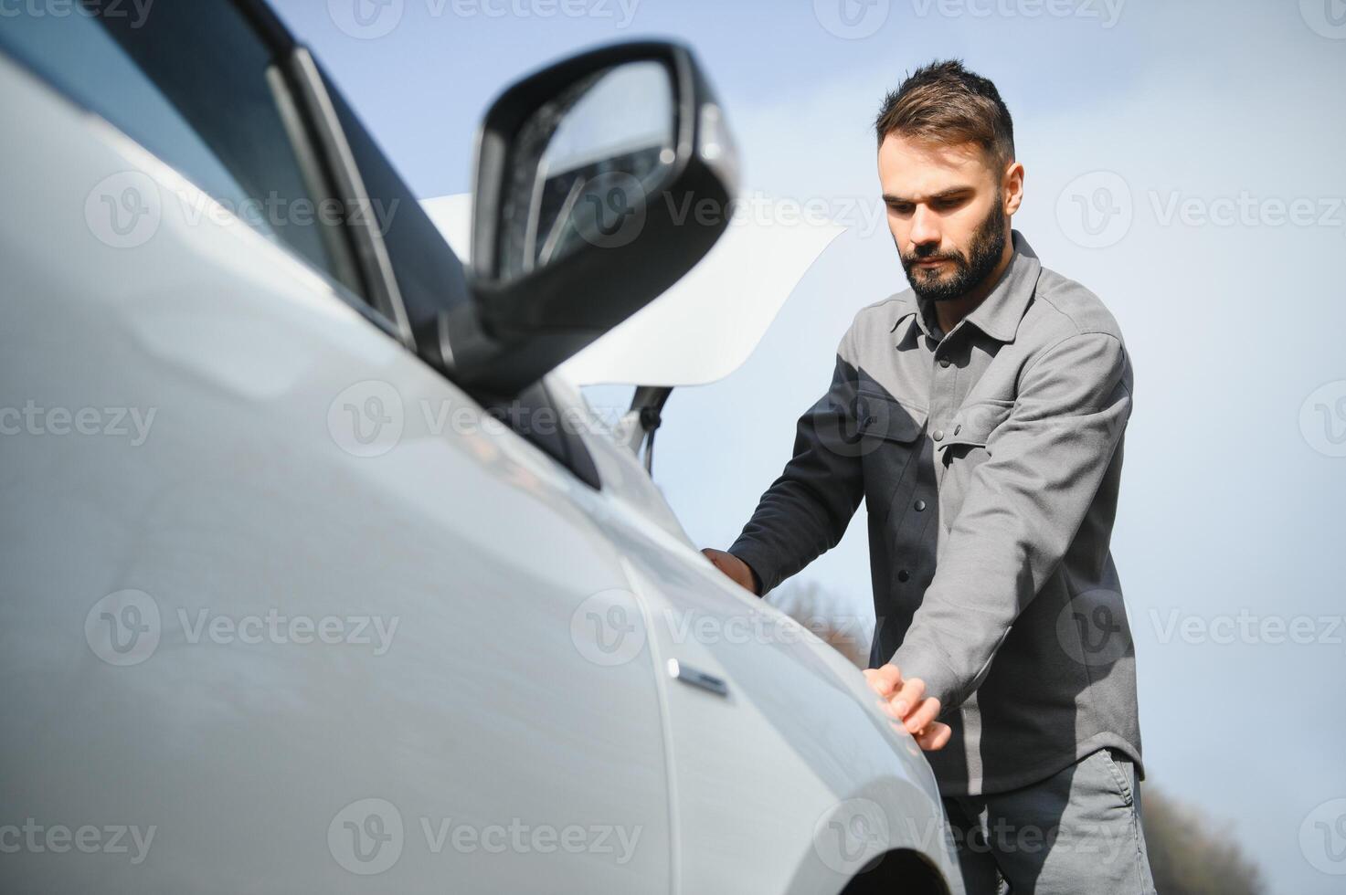 Young upset casual man trying to fix his broken car outdoors. Man waiting for towing service for help car accident on the road. Roadside assistance concept. photo