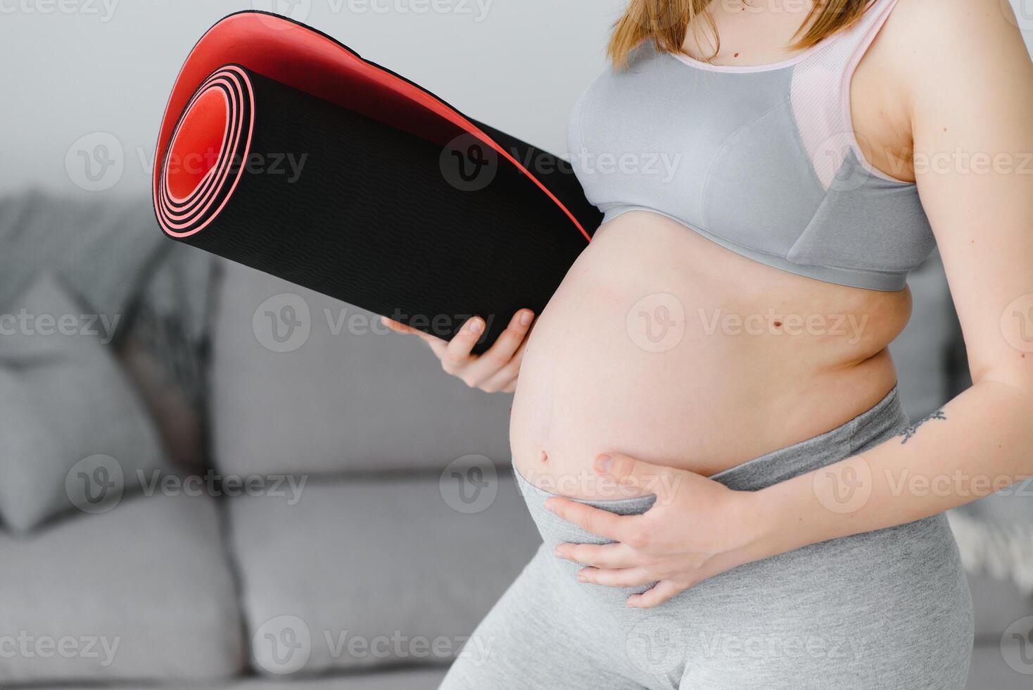 Portrait of a beautiful pregnant woman smiling and holding a rolled yoga mat under her arm. Adorable expectant mother touching her belly and preparing to start her home workout photo