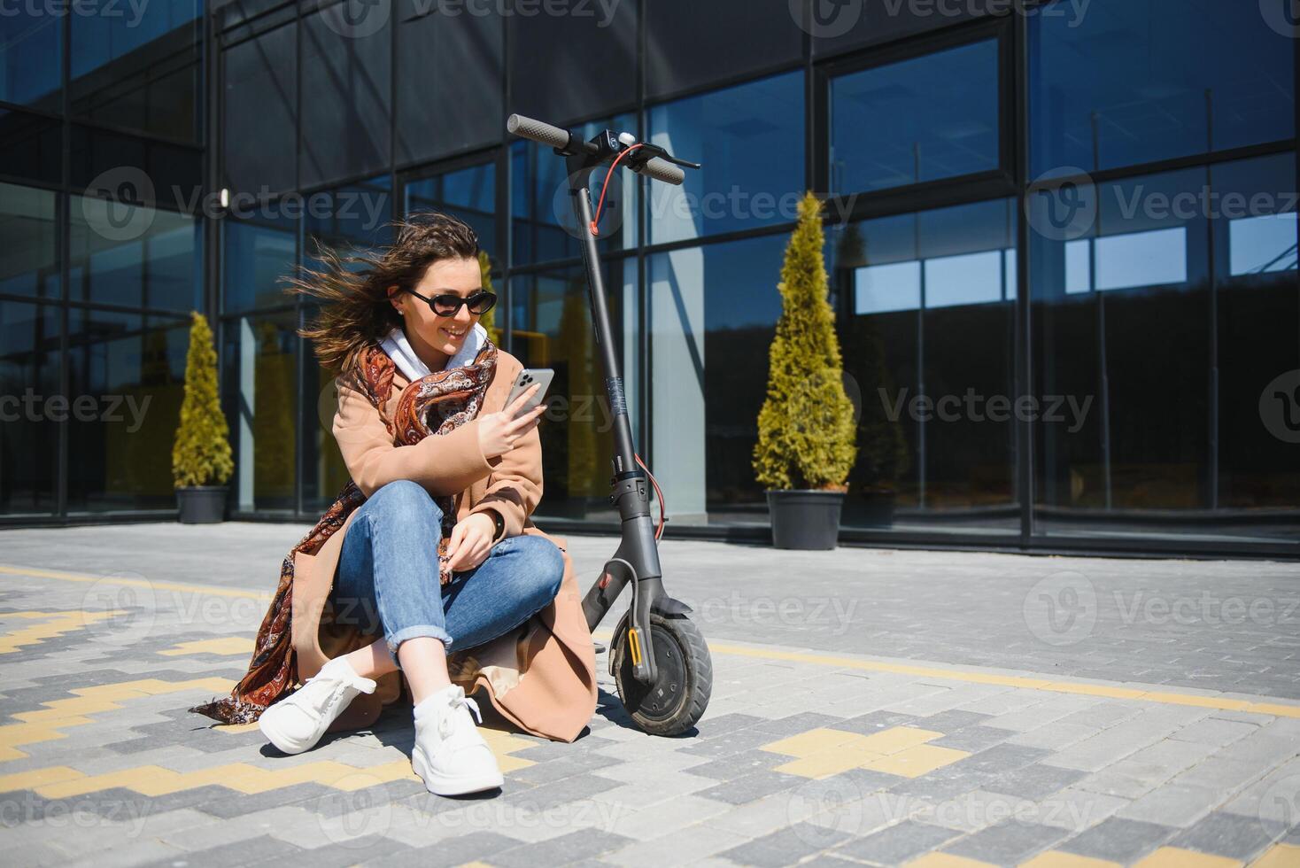 Beautiful young woman in sunglasses is sitting on her electro scooter near big glass building. photo