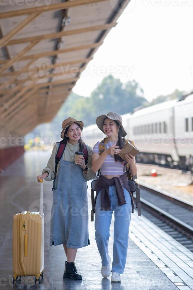 dos joven asiático amigos muchachas con mochilas a ferrocarril estación esperando para tren, dos hermosa mujer caminando a lo largo plataforma a tren estación foto