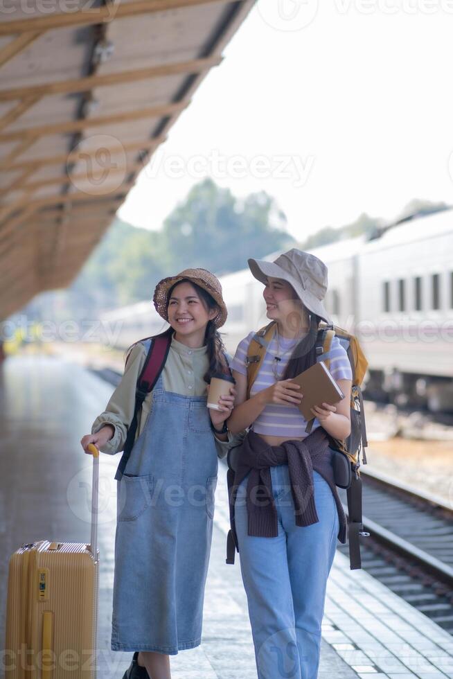 dos joven asiático amigos muchachas con mochilas a ferrocarril estación esperando para tren, dos hermosa mujer caminando a lo largo plataforma a tren estación foto