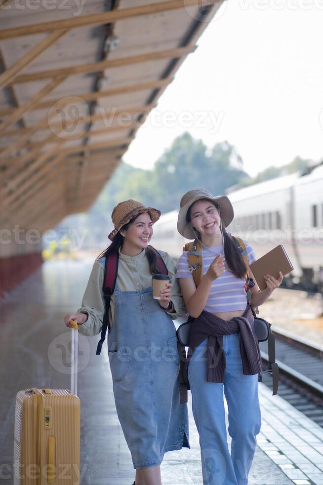 dos joven asiático amigos muchachas con mochilas a ferrocarril estación esperando para tren, dos hermosa mujer caminando a lo largo plataforma a tren estación foto