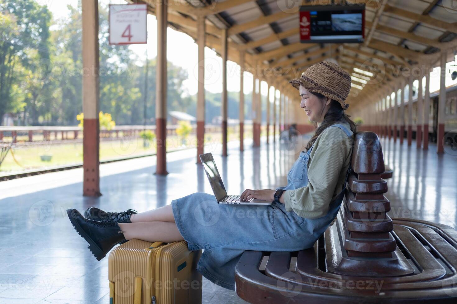 Girl using a laptop while waiting in a train station, Girl on train station with luggage working on laptop computer, laptop in use, sits with a suitcase photo